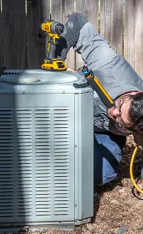 HVAC technician using a power drill to service an outdoor air conditioning unit.