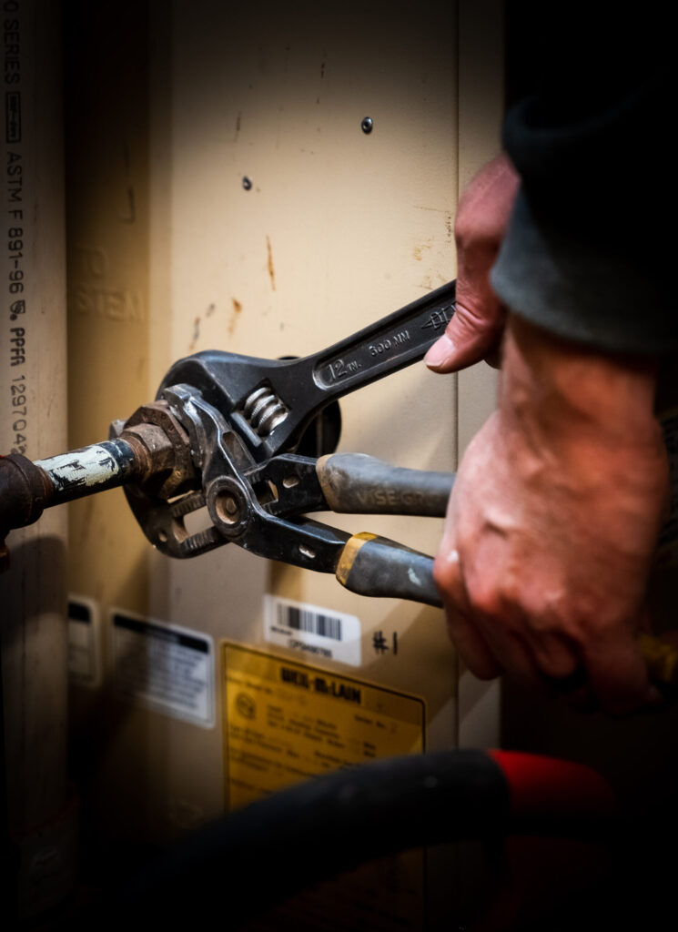 A person uses two wrenches to tighten a pipe connection on a piece of equipment with warning labels.