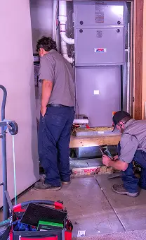 Two HVAC technicians installing or servicing a furnace unit in a utility room.