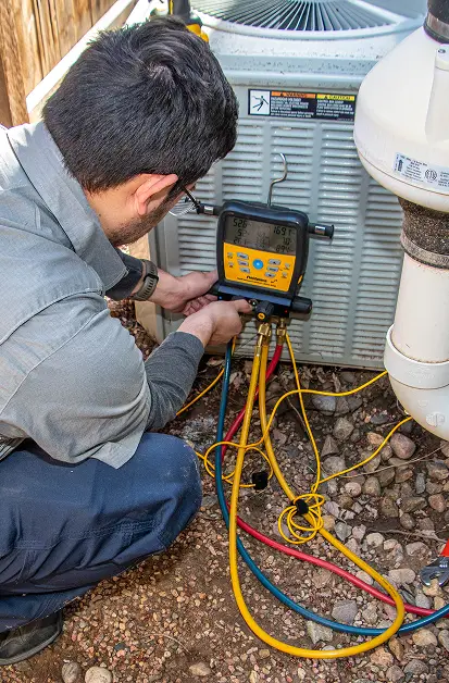HVAC technician kneeling outdoors while reading a digital manifold gauge connected to an air conditioning unit.